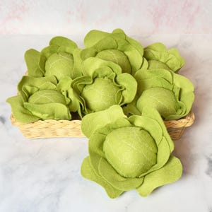 May include: A basket filled with felt green cabbage play food. The cabbages are arranged in a circular pattern with the largest cabbage in the center.