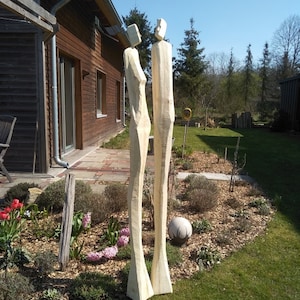 K&ouml;nnte beinhalten: Zwei hohe, abstrakte Holzskulpturen von menschlichen Figuren stehen in einem Garten. Die Skulpturen sind hell, mit einem einfachen, geometrischen Design. Der Garten hat gr&uuml;nes Gras, Blumen und eine dekorative Steinkugel.
