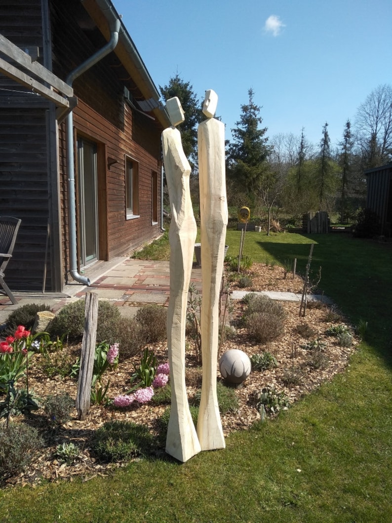 K&ouml;nnte beinhalten: Zwei hohe, abstrakte Holzskulpturen von menschlichen Figuren stehen in einem Garten. Die Skulpturen sind hell, mit einem einfachen, geometrischen Design. Der Garten hat gr&uuml;nes Gras, Blumen und eine dekorative Steinkugel.