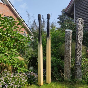 Könnte beinhalten: Zwei große Holzskulpturen in Form von Streichhölzern stehen in einem Garten. Die Köpfe sind schwarz verkohlt, die Schäfte zeigen Naturholz. Die Skulpturen stehen vor grünem Laub und blauem Himmel.