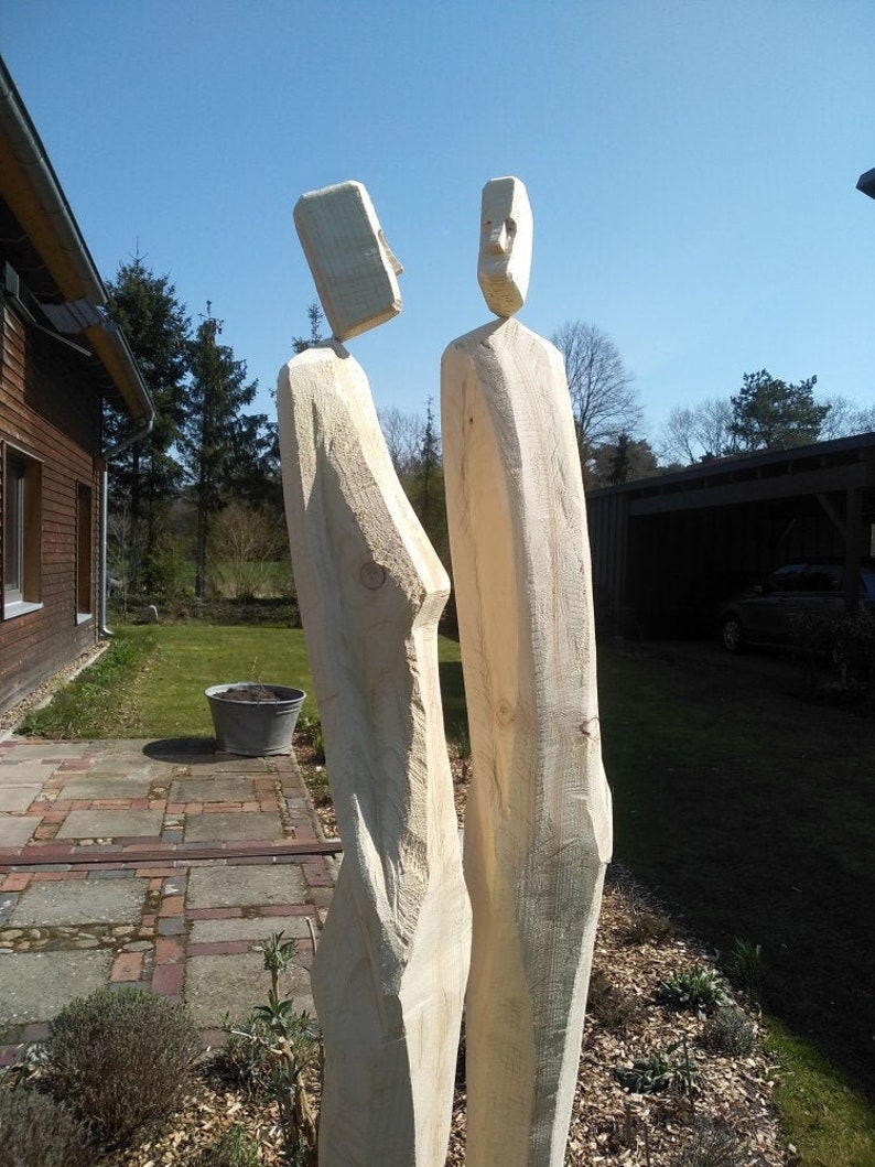 May include: Two wooden sculptures of people standing side-by-side. The sculptures are carved from a single piece of wood and have a simple, abstract style.