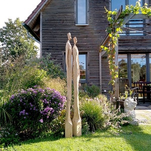 K&ouml;nnte beinhalten: Zwei hohe Holzskulpturen von menschlichen Figuren stehen in einem Garten. Die Skulpturen sind hellbraun und haben ein einfaches, abstraktes Design. Der Garten hat gr&uuml;nes Gras, lila Blumen und ein Holzhaus im Hintergrund.