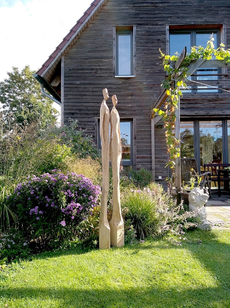 K&ouml;nnte beinhalten: Zwei hohe Holzskulpturen von menschlichen Figuren stehen in einem Garten. Die Skulpturen sind hellbraun und haben ein einfaches, abstraktes Design. Der Garten hat gr&uuml;nes Gras, lila Blumen und ein Holzhaus im Hintergrund.
