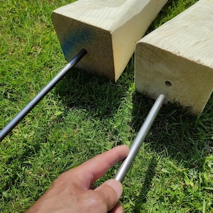 May include: Two wooden posts with metal rods inserted through holes in the top. The posts are on a bed of green grass.