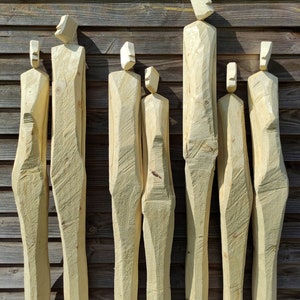May include: Seven wooden sculptures of human figures, each carved from a single piece of wood, standing in a row against a wood panel background.