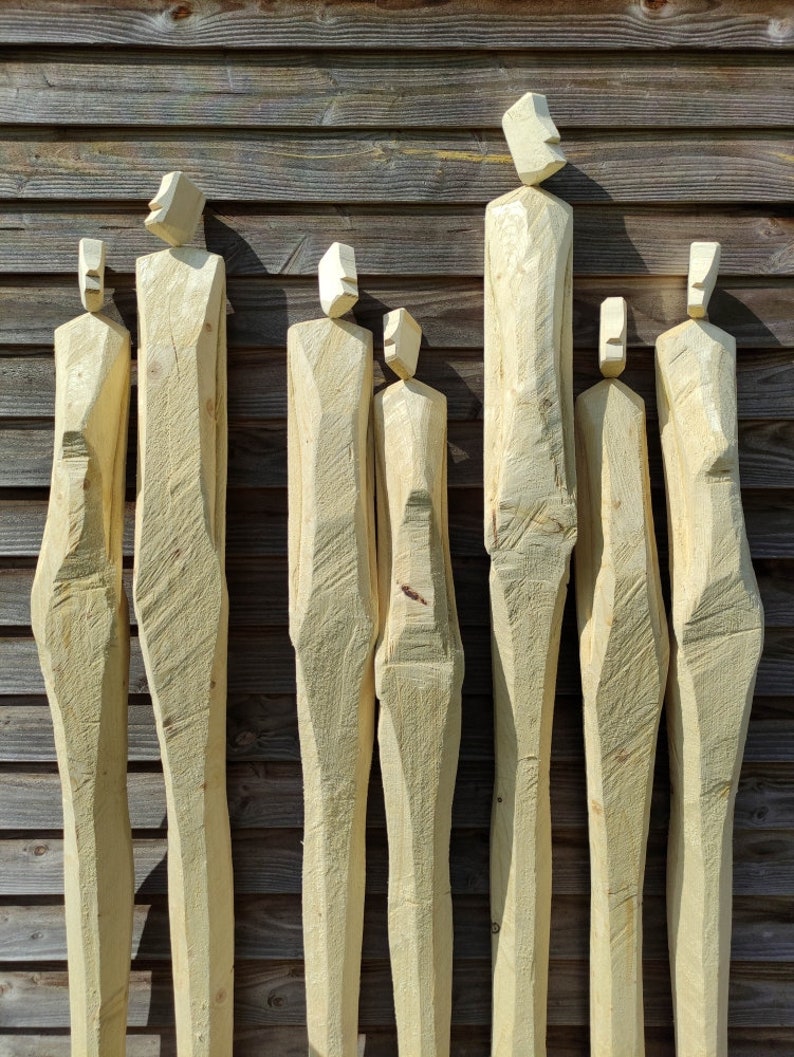May include: Seven wooden sculptures of human figures, each carved from a single piece of wood, standing in a row against a wood panel background.
