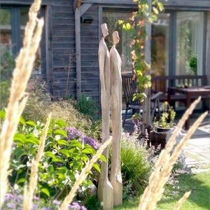 May include: A wooden sculpture of two figures standing side-by-side in a garden setting. The figures are carved from a single piece of wood and are painted in a natural wood tone.