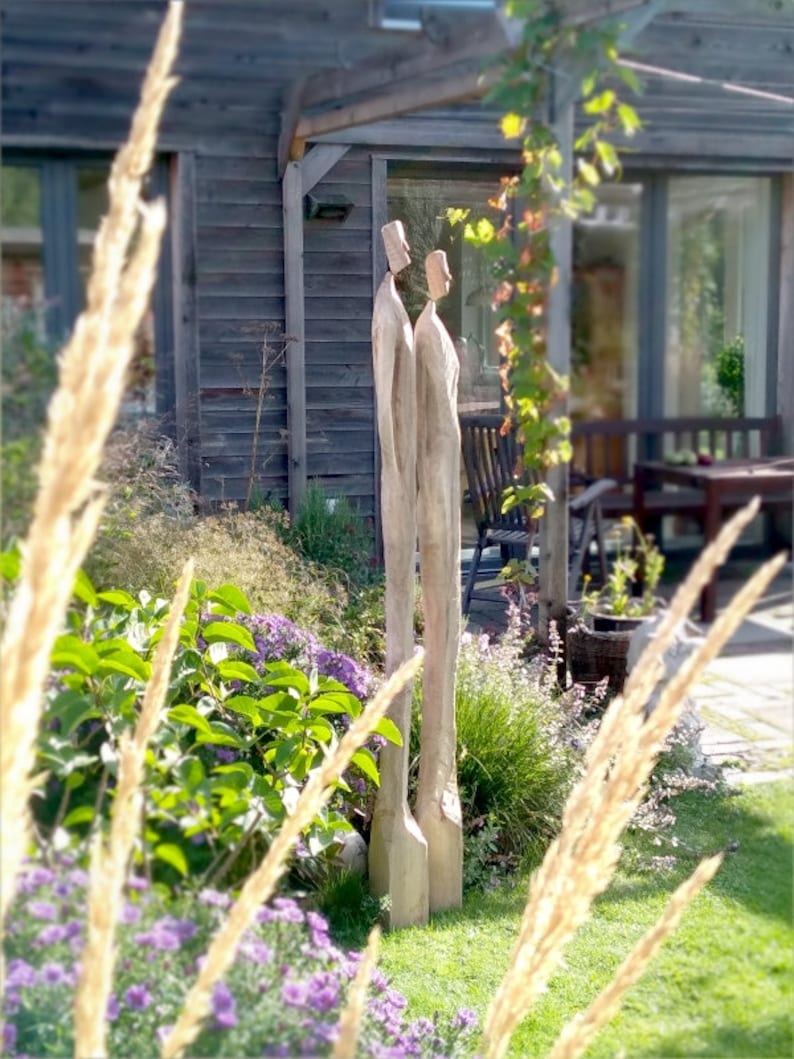 May include: A wooden sculpture of two figures standing side-by-side in a garden setting. The figures are carved from a single piece of wood and are painted in a natural wood tone.