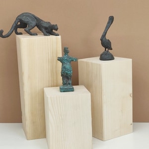 Könnte beinhalten: Drei Holz-Präsentationsständer mit drei Bronzeskulpturen darauf. Die Skulpturen sind ein Panther, ein Pelikan und eine menschliche Figur. Die Ständer sind alle gleich groß und aus hellem Holz.