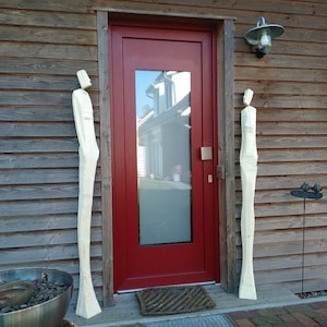 May include: Two tall, white wooden sculptures stand on either side of a red door with a glass pane. The door is set into a wooden wall with horizontal siding.