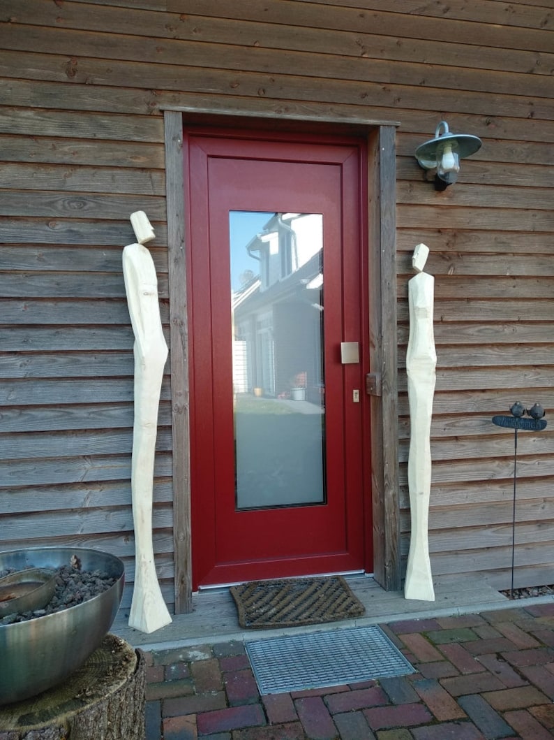 May include: Two tall, white wooden sculptures stand on either side of a red door with a glass pane. The door is set into a wooden wall with horizontal siding.