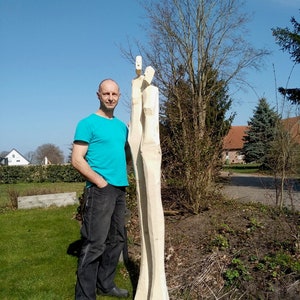 May include: A man stands next to two tall, wooden sculptures of people. The sculptures are carved from a single piece of wood and are very detailed. The man is wearing a blue shirt and jeans. The sculptures are in a garden setting with green grass and trees in the background.