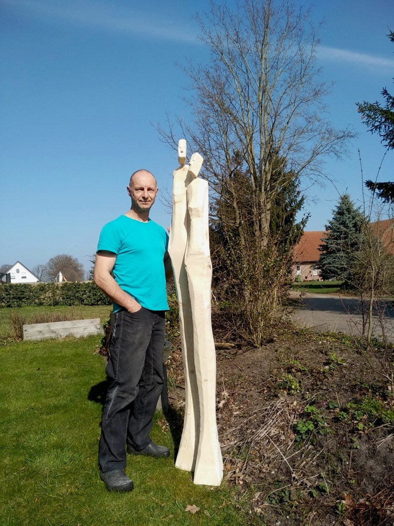 May include: A man stands next to two tall, wooden sculptures of people. The sculptures are carved from a single piece of wood and are very detailed. The man is wearing a blue shirt and jeans. The sculptures are in a garden setting with green grass and trees in the background.