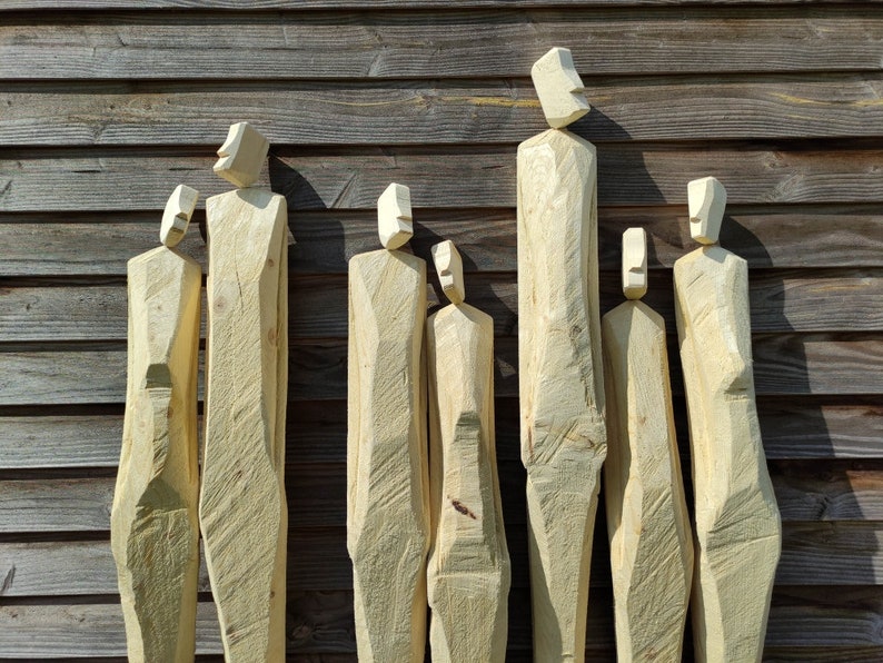 May include: Seven wooden sculptures of abstract human figures, carved from light brown wood, stand in a row against a wood panel background.