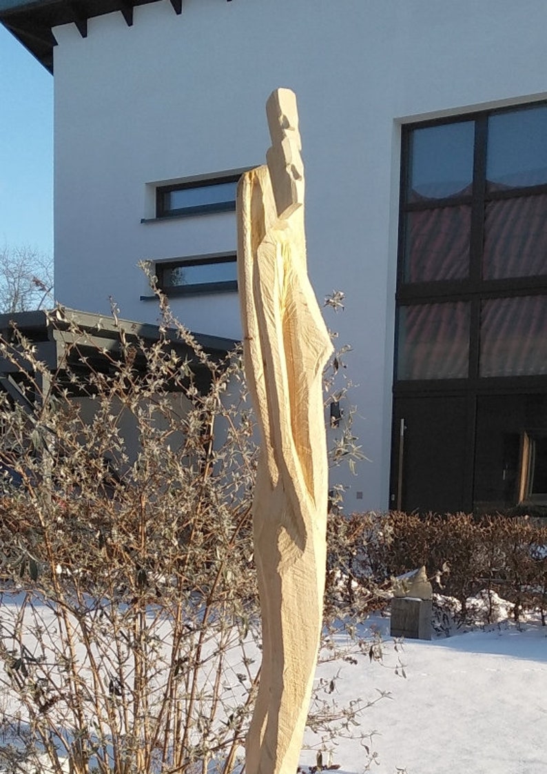 May include: A wooden sculpture of a person standing in front of a building with a large window. The sculpture is carved in a minimalist style and is made of light-colored wood.