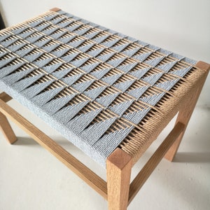 Puede incluir: Un taburete de madera con un asiento tejido en color azul claro y natural. El asiento está tejido en forma de diamante.