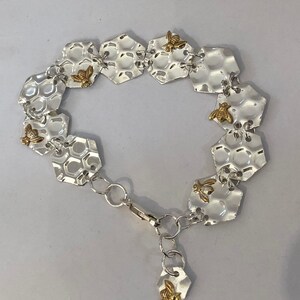 May include: A silver bracelet featuring a honeycomb design and gold bee charms. The bracelet is made up of multiple hexagonal links joined by small silver chains. The bee charms are attached to the links and have a textured surface.