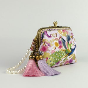 May include: A small, purple and white purse with a gold frame and a peacock design. The purse has a pearl chain strap and two pink and purple tassels.