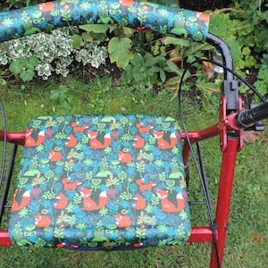 以下が含まれることがあります： 赤い歩行器に黒いパッド入りのハンドルと黒い座面があり、座面にはキツネのプリントが施されています。座面はキツネ、花、葉の模様の生地で覆われています。