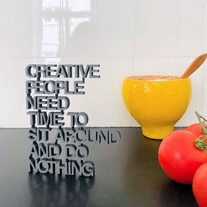 Peut inclure: Une sculpture en 3D de mots gris qui dit "Creative People Need Time To Sit Around And Do Nothing". La sculpture est sur une surface noire avec un bol jaune et des tomates rouges.