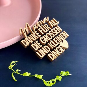 Peut inclure: Une pancarte en bois avec le texte "Mama Danke fur all die grossen und kleinen dinge" avec un petit cœur à la fin. La pancarte est sur une surface noire.