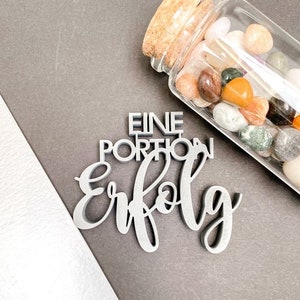 Puede incluir: Un letrero de madera plateado con el texto "Eine Portion Erfolg" que se traduce como "Una porción de éxito". El letrero está sobre una superficie gris junto a un frasco de vidrio lleno de piedras de colores.
