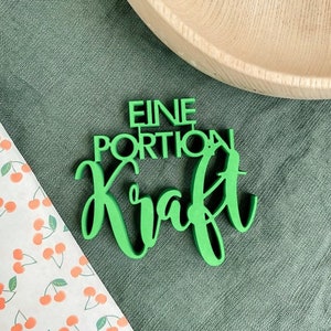 Puede incluir: Un letrero de madera verde con el texto "EINE PORTION KRAFT" en una fuente decorativa.