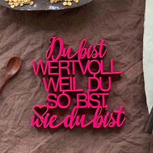 Op de afbeelding: Roze houten bord met de tekst "Du bist wertvoll weil du so bist wie du bist", wat vertaald wordt als "Je bent waardevol omdat je bent zoals je bent".
