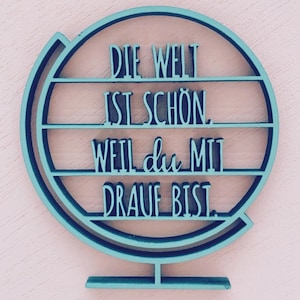 Könnte beinhalten: Ein tealblauer Holzglobus mit dem Text "Die Welt ist schön, weil du mit drauf bist." Der Globus befindet sich auf einem rosafarbenen Hintergrund.