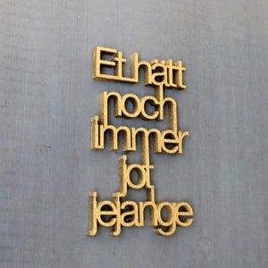 Peut inclure: Lettres en bois de couleur dorée épellant la phrase "Et hatt noch immer jot jelange" sur un fond gris.