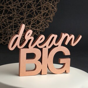 Puede incluir: Un letrero de madera de color cobre con las palabras "dream big" en una fuente cursiva.