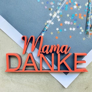 Può includere: Un cartello in legno rosso con le parole "Mama Danke" in un font corsivo. Il cartello è su una superficie bianca con coriandoli colorati sparsi intorno.