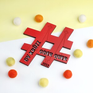Könnte beinhalten: Rotes Holz-Hashtag-Schild mit dem Text "OV KRÜTZ ODER QUER 2023" in Weiß. Das Schild ist von bunten Pompons umgeben.