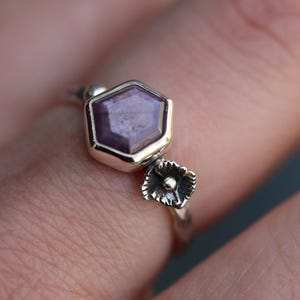 Könnte beinhalten: Ein silberner Ring mit einem sechseckigen violetten Edelstein und einem kleinen Blumendesign.