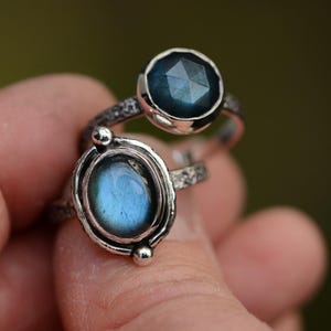 Könnte beinhalten: Zwei silberne Ringe mit blauen Edelsteinen. Ein Ring hat einen runden, facettierten Stein, während der andere einen oval geformten, glatten Stein hat. Die Ringe werden vor einem verschwommenen grünen Hintergrund gehalten.