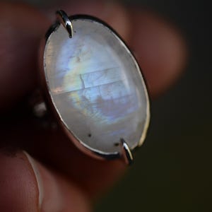 Könnte beinhalten: Ein Silberring mit einem großen, ovalen Mondstein-Cabochon, der milchig-transluzent erscheint und irisierende blaue sowie regenbogenfarbene Lichtreflexe aufweist. Der Edelstein ist in einer einfachen Metallfassung mit zwei sichtbaren Krappen befestigt.