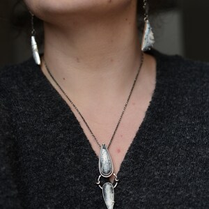 May include: A silver necklace with a teardrop-shaped pendant featuring a white and gray stone. The pendant is attached to a smaller, pointed pendant with a similar stone. The necklace is worn by a person with a gray sweater.