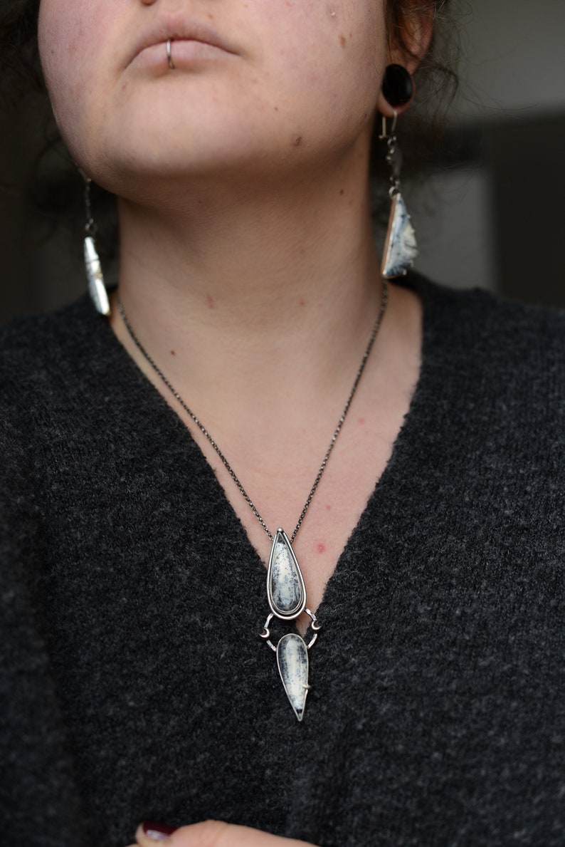 May include: A silver necklace with a teardrop-shaped pendant featuring a white and gray stone. The pendant is attached to a smaller, pointed pendant with a similar stone. The necklace is worn by a person with a gray sweater.