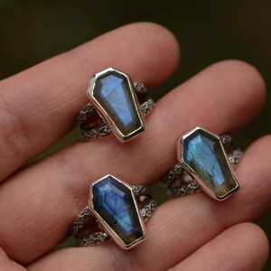 May include: Three silver rings with coffin-shaped labradorite stones. The stones display iridescent blue and gold hues. The rings have a textured band design, and are displayed on a hand.