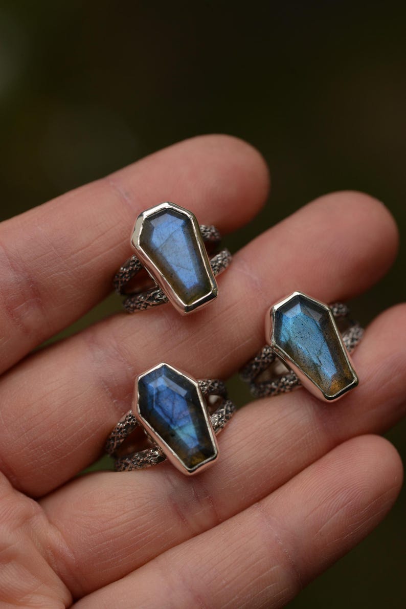 May include: Three silver rings with coffin-shaped labradorite stones. The stones display iridescent blue and gold hues. The rings have a textured band design, and are displayed on a hand.