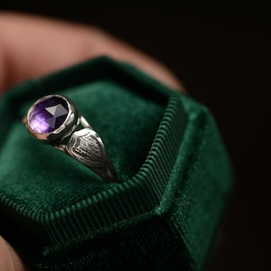 May include: A silver ring with a purple gemstone set in a bezel setting. The ring is in a green velvet ring box.