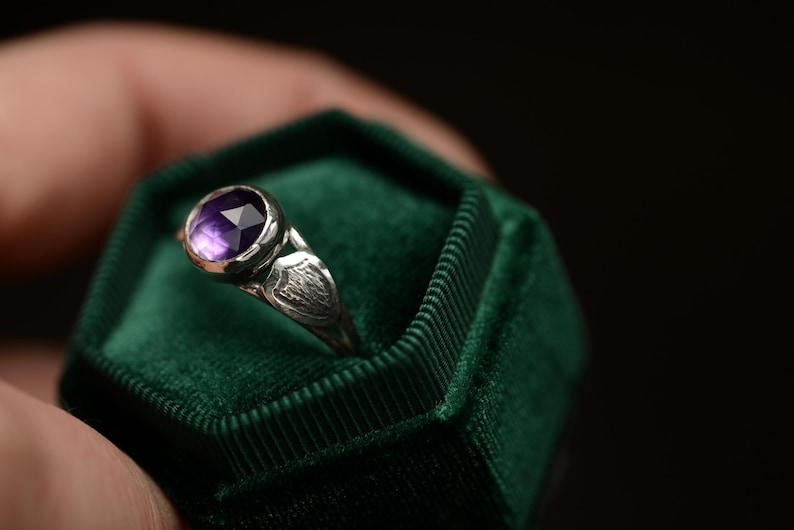 May include: A silver ring with a purple gemstone set in a bezel setting. The ring is in a green velvet ring box.