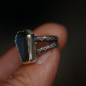 May include: A silver ring with a coffin-shaped, blue-green gemstone. The ring has a double band with a textured pattern. The gemstone is set in a silver bezel, creating a striking contrast.