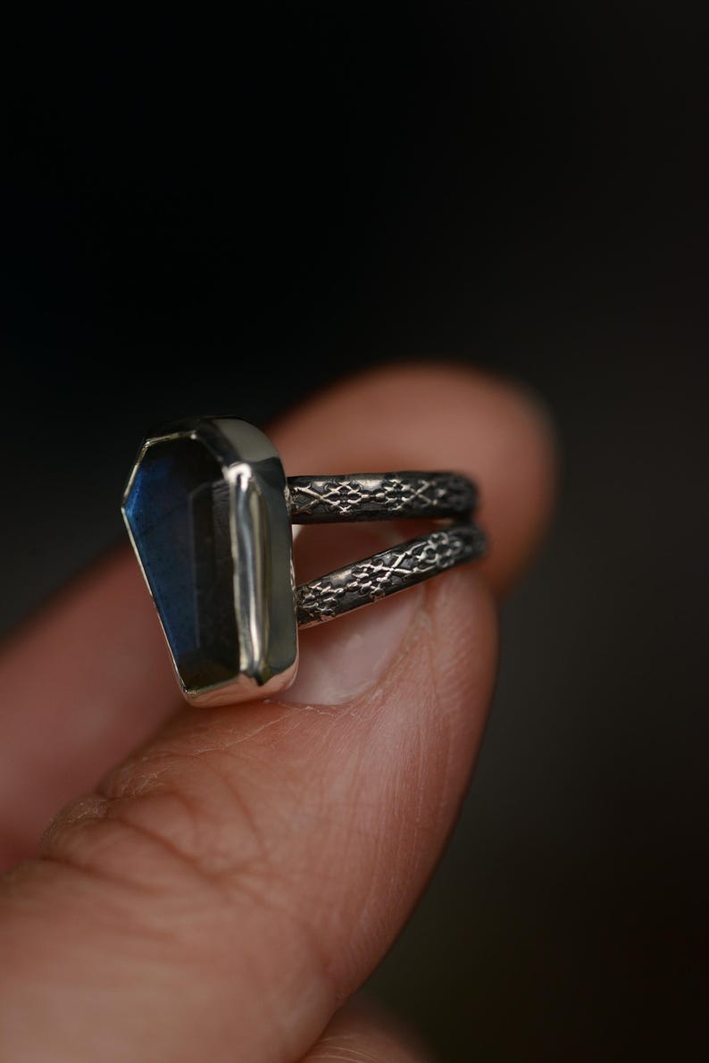 May include: A silver ring with a coffin-shaped, blue-green gemstone. The ring has a double band with a textured pattern. The gemstone is set in a silver bezel, creating a striking contrast.