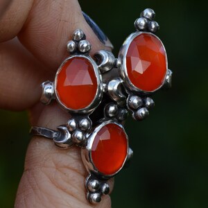 Könnte beinhalten: Ein Silberring mit drei ovalen, facettierten, orangeroten Edelsteinen. Der Ring hat ein strukturiertes Band und dekorative Silberakzente um die Edelsteine. Der Ring wird vor einem verschwommenen grünen Hintergrund gehalten.