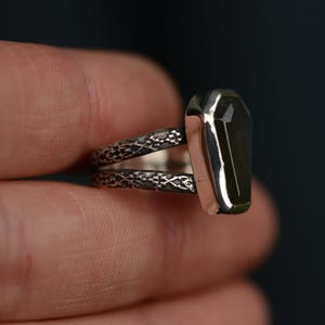 May include: A silver ring with a coffin-shaped, dark green stone. The ring has a split band with an ornate, textured design. The stone is set in a polished silver bezel.