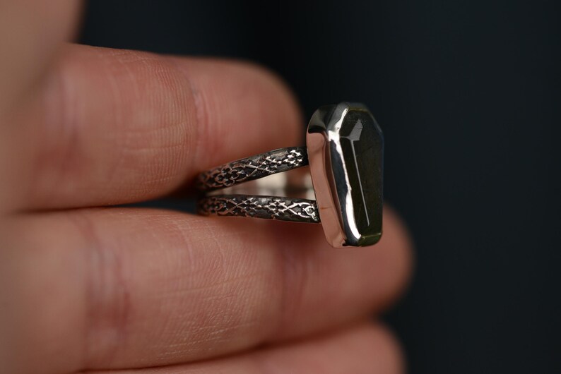 May include: A silver ring with a coffin-shaped, dark green stone. The ring has a split band with an ornate, textured design. The stone is set in a polished silver bezel.