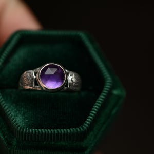 May include: A silver ring with a purple amethyst gemstone set in a bezel setting. The ring is in a green velvet ring box.