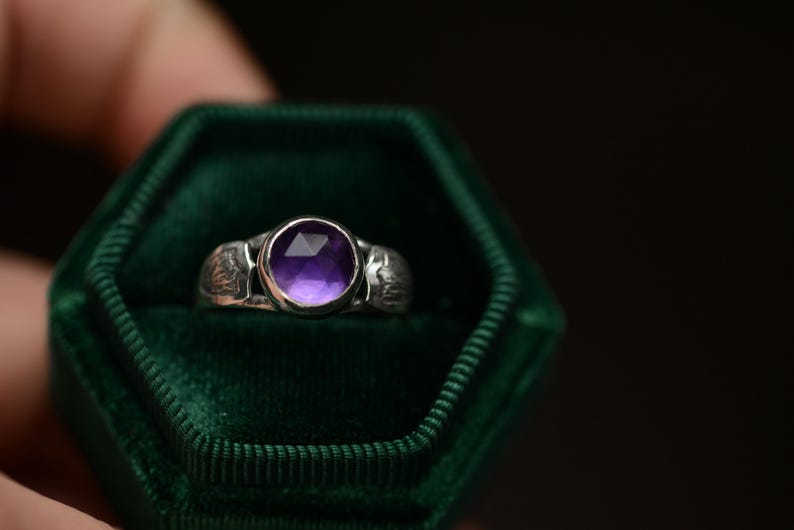 May include: A silver ring with a purple amethyst gemstone set in a bezel setting. The ring is in a green velvet ring box.