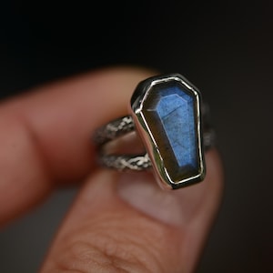May include: A silver ring featuring a coffin-shaped, blue-hued gemstone. The gemstone is set in a silver bezel, and the band has a textured, patterned design. The ring is held up against a dark background.